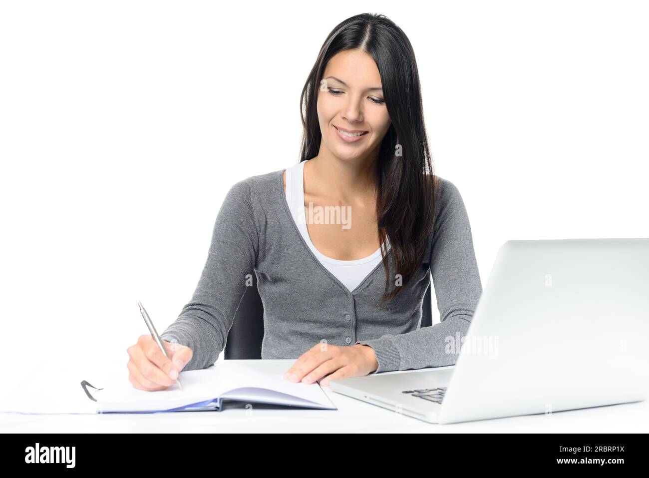 Giovane donna sorridente che lavora a una scrivania con il suo computer portatile scrivendo appunti in un notebook concettuali di un lavoratore d'ufficio laborioso o Foto Stock