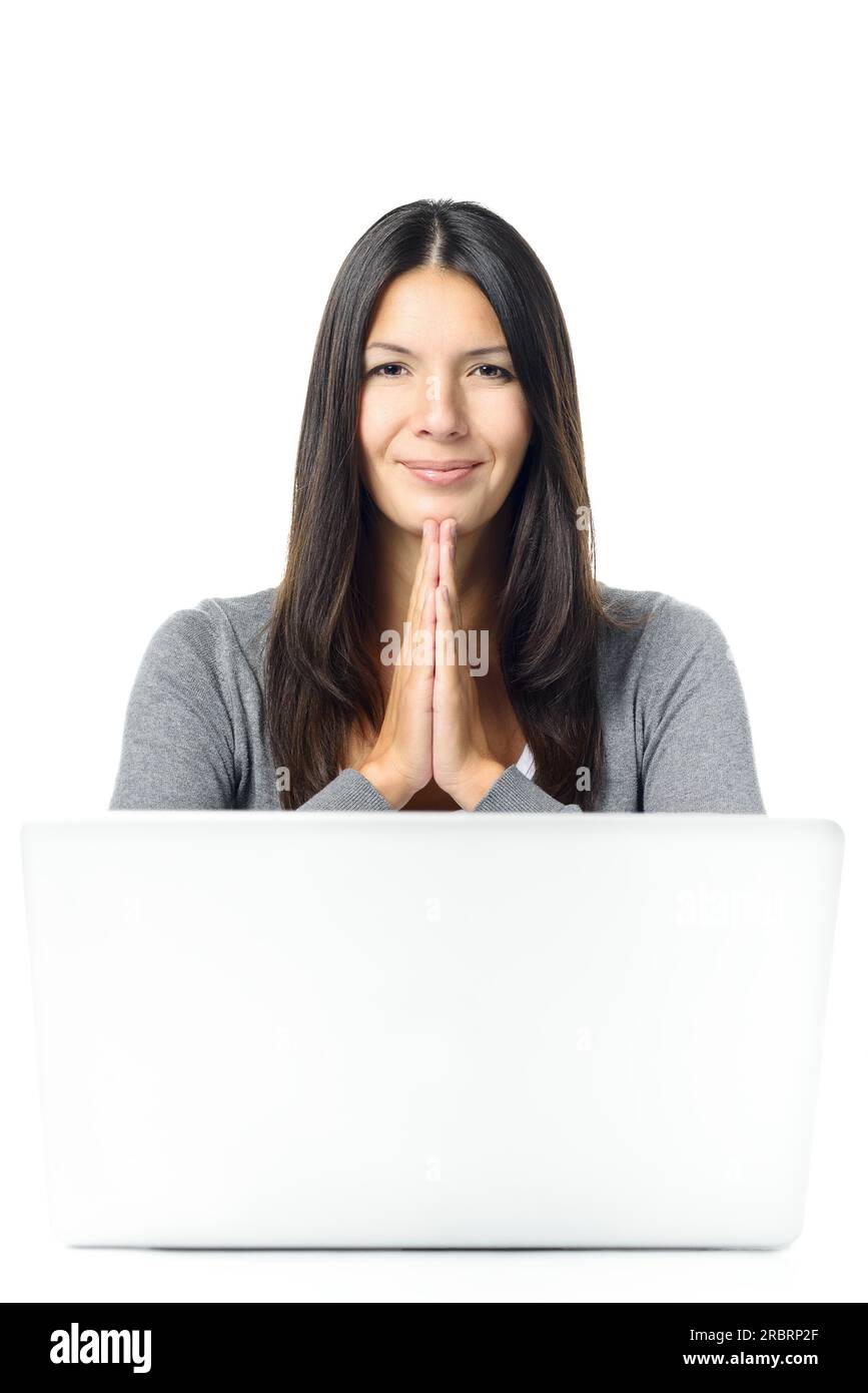 Giovane studentessa o donna d'affari con un sorriso e mani stringute in gratitudine o preghiera seduta alla sua scrivania dietro un computer portatile Foto Stock