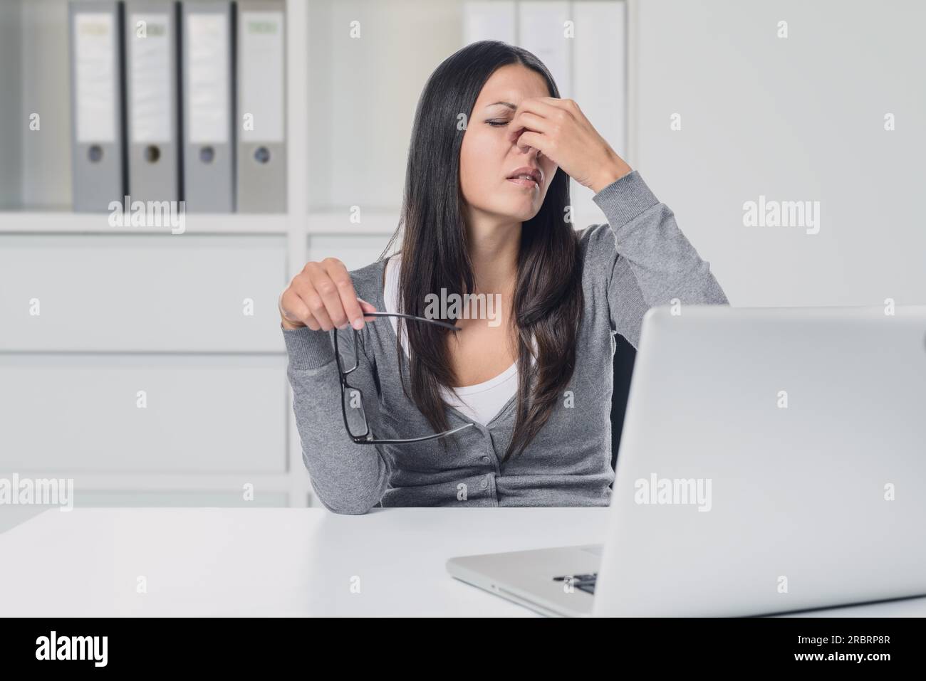 Una giovane donna affaticata dagli occhi al suo laptop che si rimuove gli occhiali per strofinarsi gli occhi con il dito con un'espressione dolorosa Foto Stock