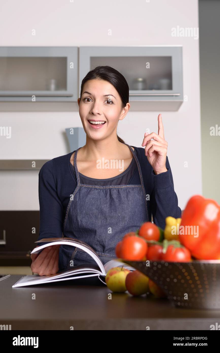 Felice donna di mezza età che indossa grembiule al bancone della cucina con ricettario, enfatizzando il segno positivo della mano Foto Stock