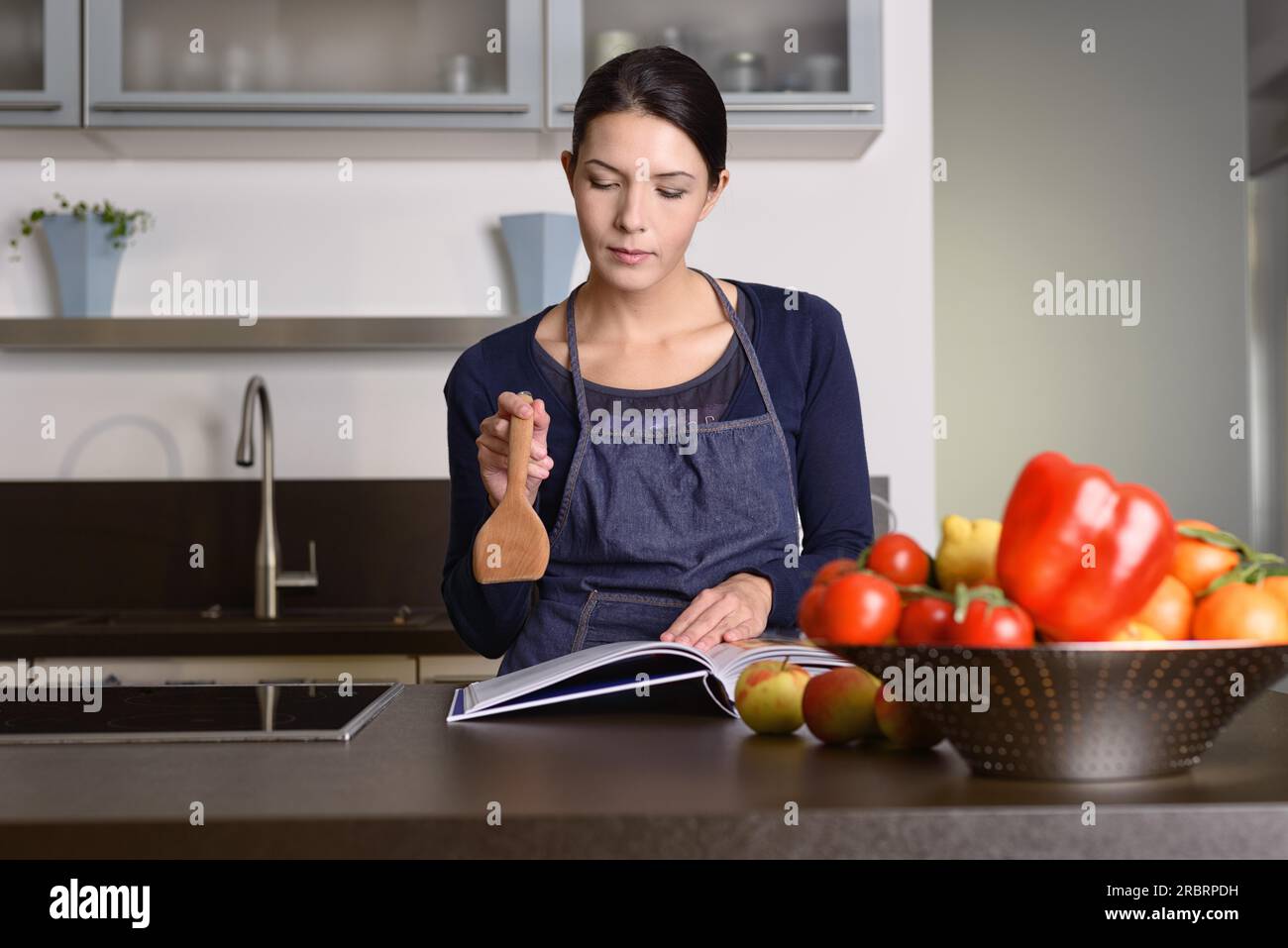 Una bella donna seria che indossa un grembiule che tiene in mano una Ladle di legno mentre legge un ricettario al tavolo con frutta e verdure in cucina Foto Stock