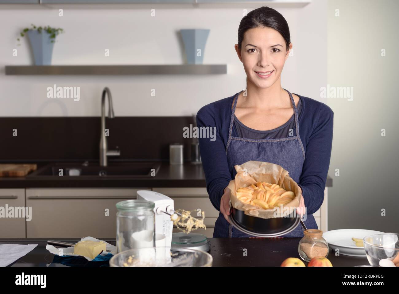 Una giovane donna sorridente che fa la cottura in cucina, con orgoglio, presenta la sua fresca crostata di mele su un bancone con mele fresche Foto Stock