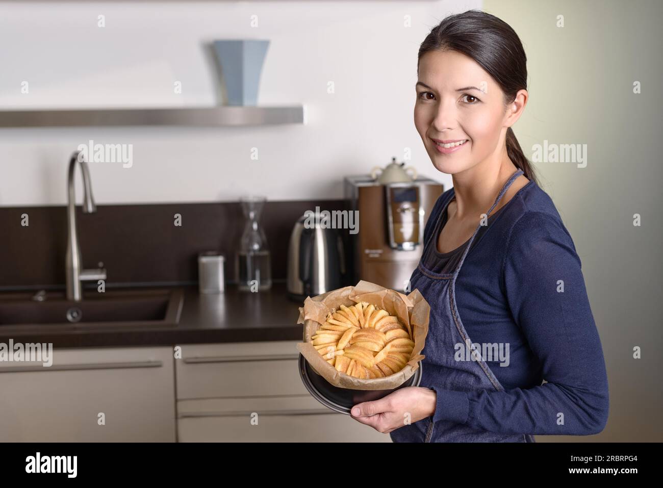 Una giovane donna sorridente che fa la cottura in cucina, con orgoglio, presenta la sua fresca crostata di mele su un bancone con mele fresche Foto Stock