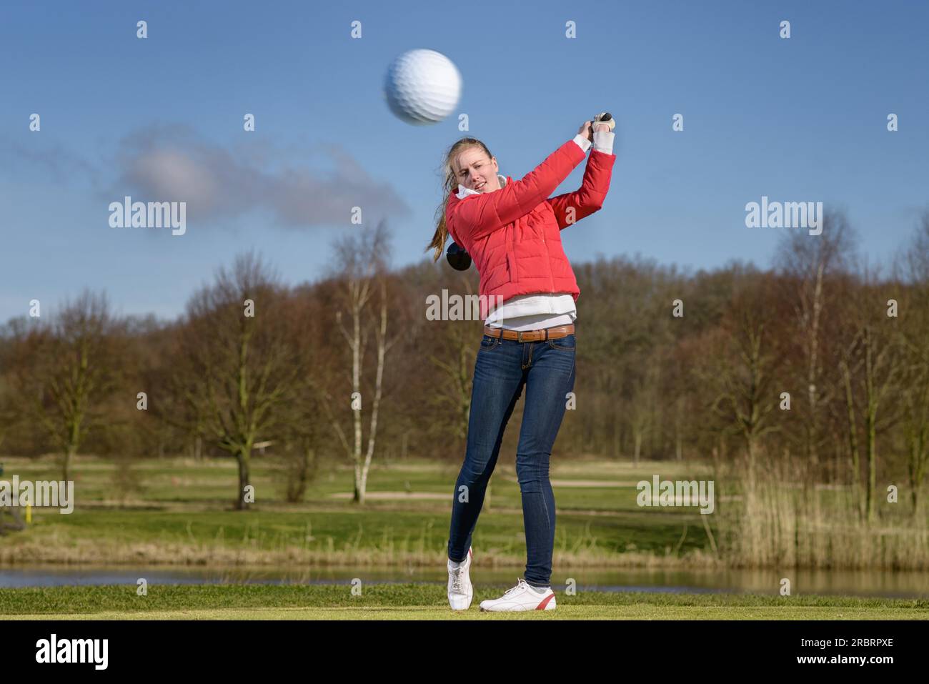 Donna golfista che colpisce la palla da golf con un autista di fronte a un pericolo d'acqua su un campo da golf con la palla che vola in aria verso la telecamera Foto Stock
