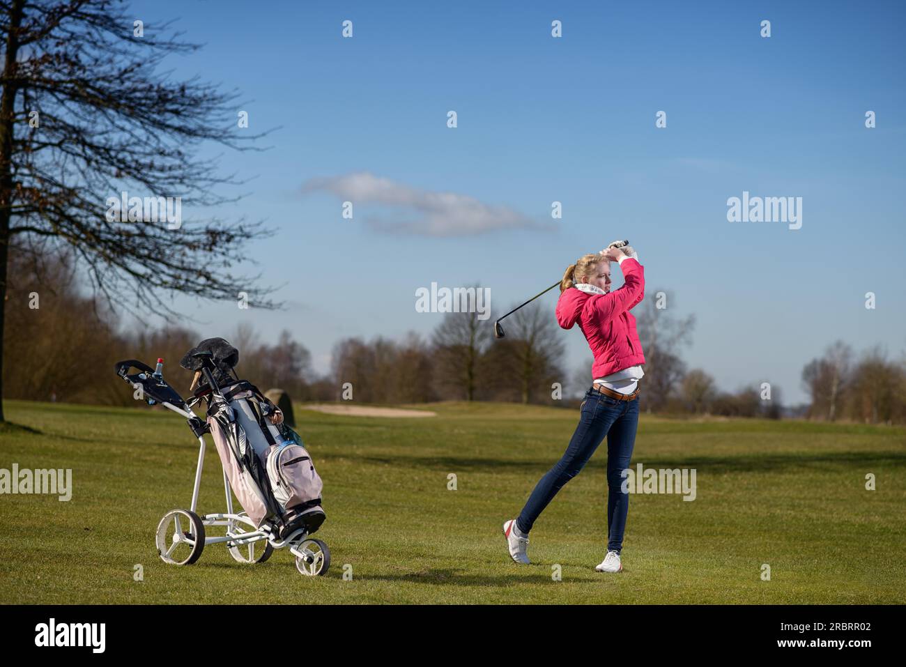 Attraente e snella giovane golfista che gioca a un tiro di avvicinamento sul fairway guardando il volo della sua palla dopo il colpo con la sua borsa da golf Foto Stock