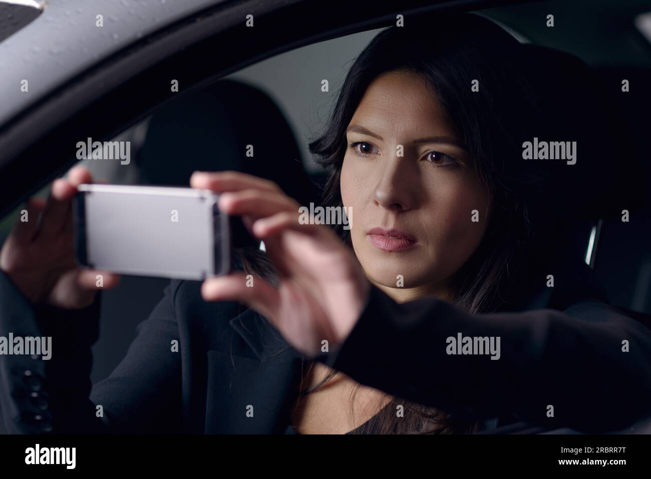 Donna attraente e seria che scatta un selfie sul cellulare mentre aspetta in auto al buio con il volto illuminato dalla luce dello schermo Foto Stock