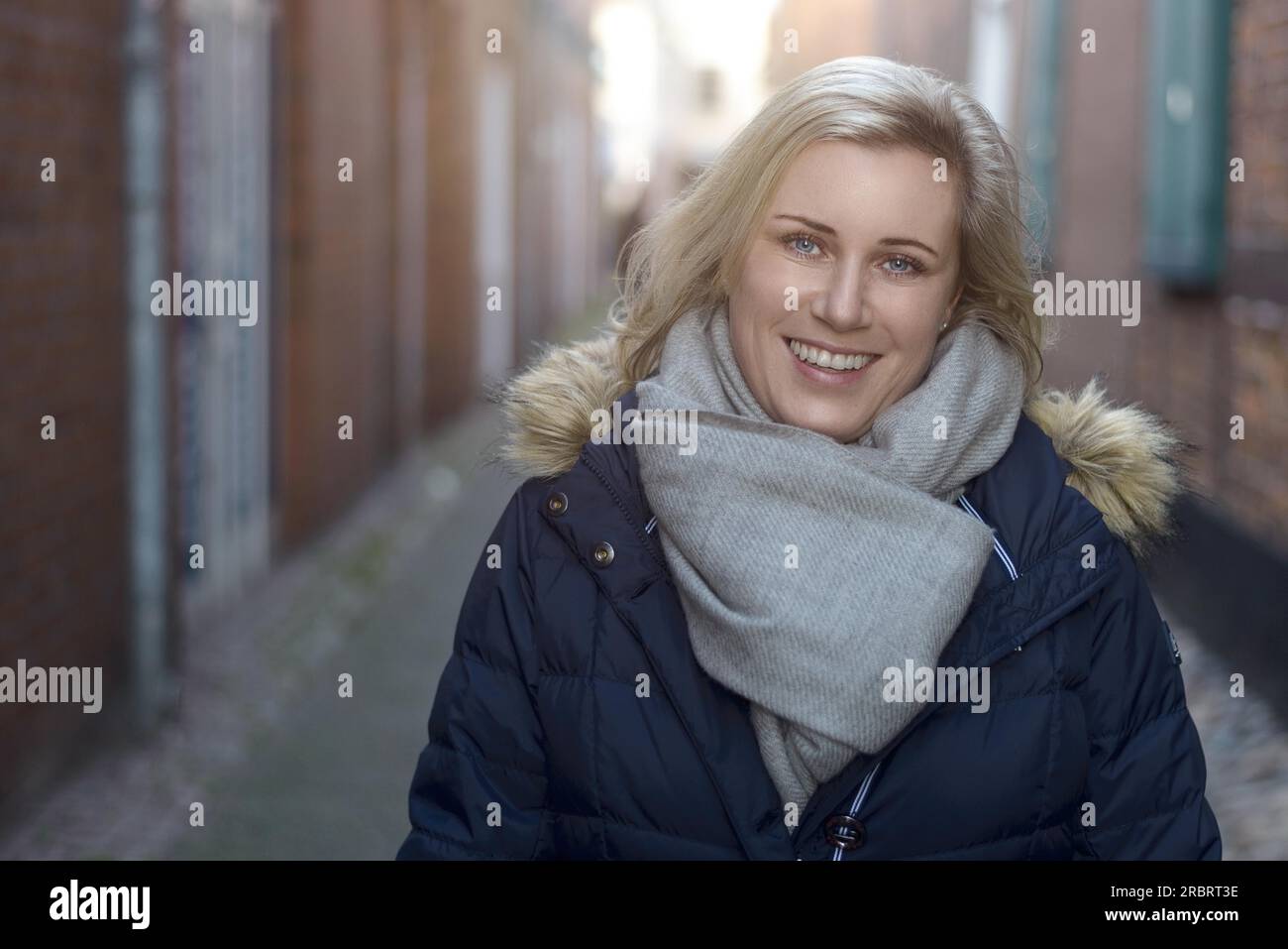 Attraente friendly donna bionda che indossa un elegante sciarpa invernale in piedi in una stretta viuzza in città sorridente felicemente con la videocamera Foto Stock