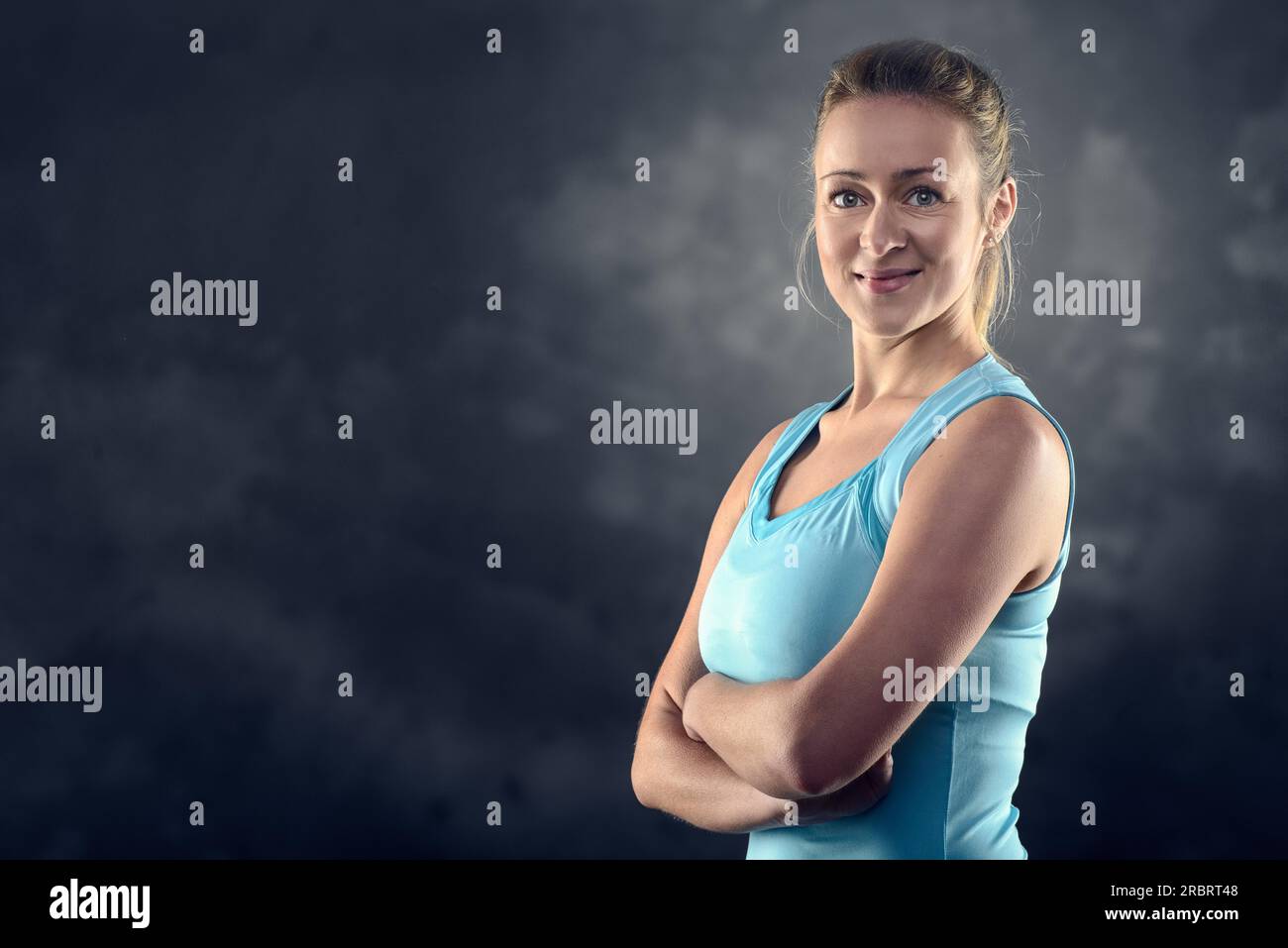 Testa e spalle Ritratto di una donna bionda atletica che indossa una canotta turchese e si trova in piedi con le braccia incrociate in Studio con sfondo grigio Foto Stock