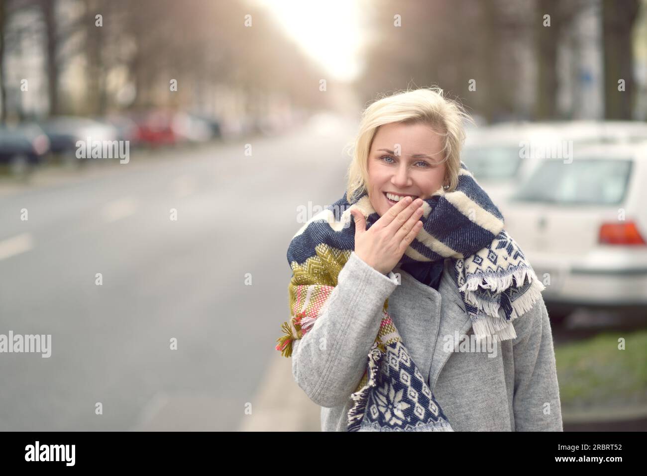 Sorridente giovane donna bionda con una sciarpa lanosa in piedi sul lato di una strada urbana guardando la telecamera e la parte superiore del corpo Foto Stock