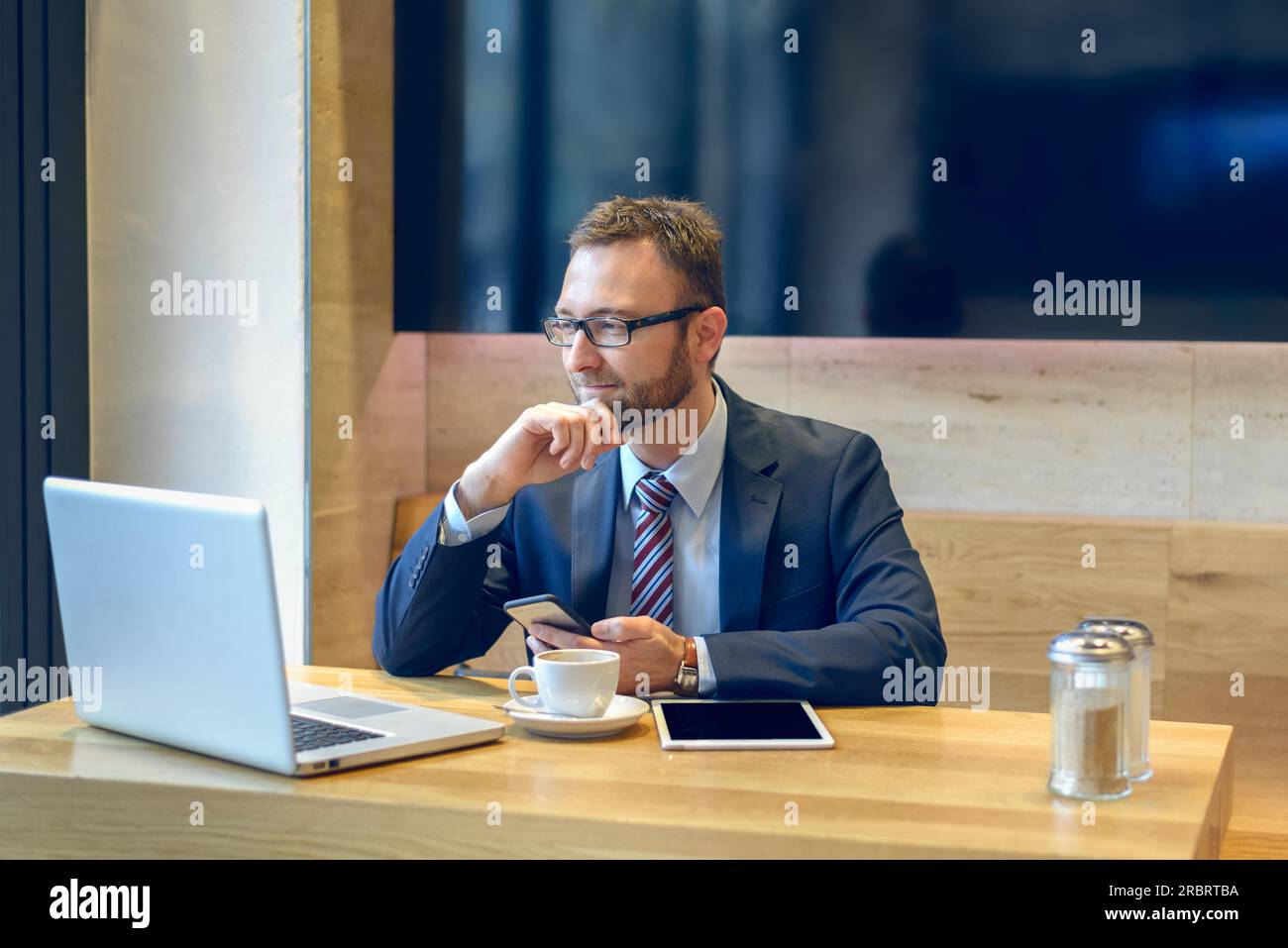 Imprenditore godendo di caffè e controllare il suo telefono cellulare per i messaggi con un sorriso con il suo computer portatile e tablet aperto sulla tavola Foto Stock