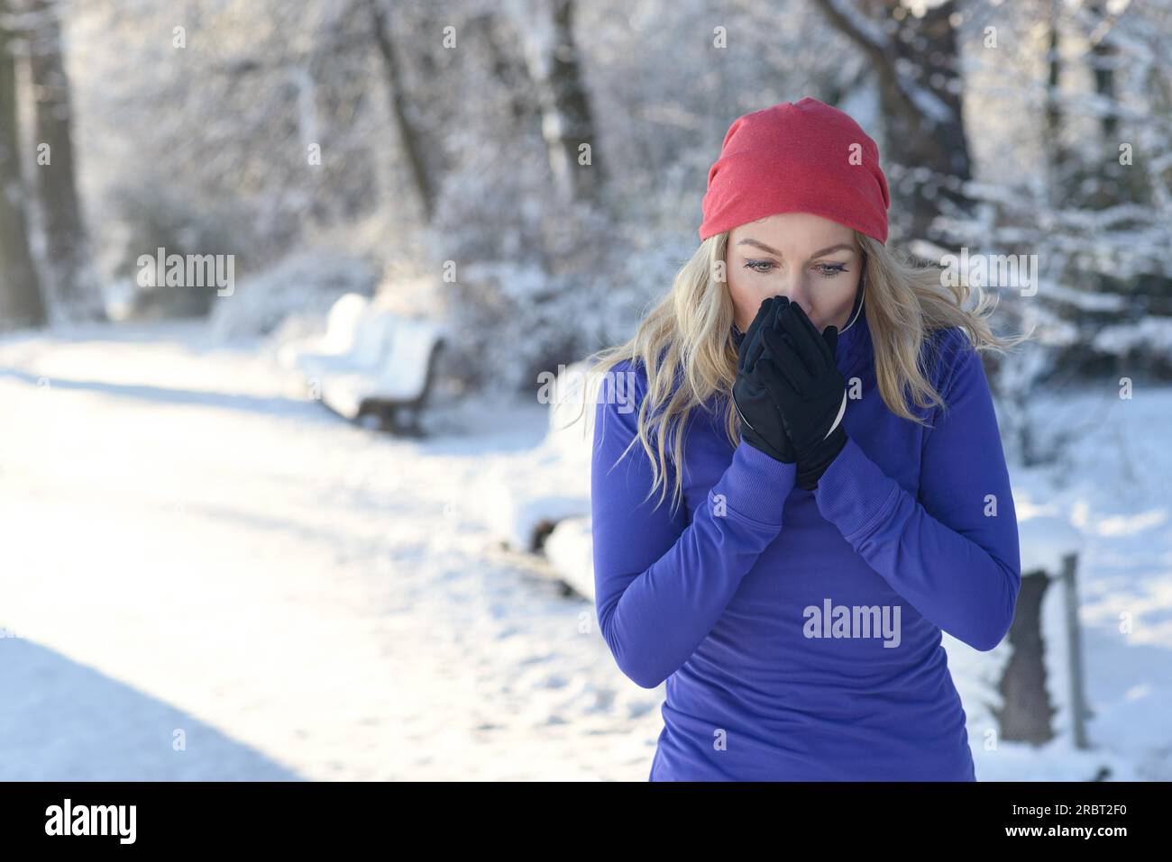 Bella giovane donna bionda che soffia sulle mani dei guanti per tenerli al caldo in un freddo parco invernale sulla neve al sole del mattino Foto Stock