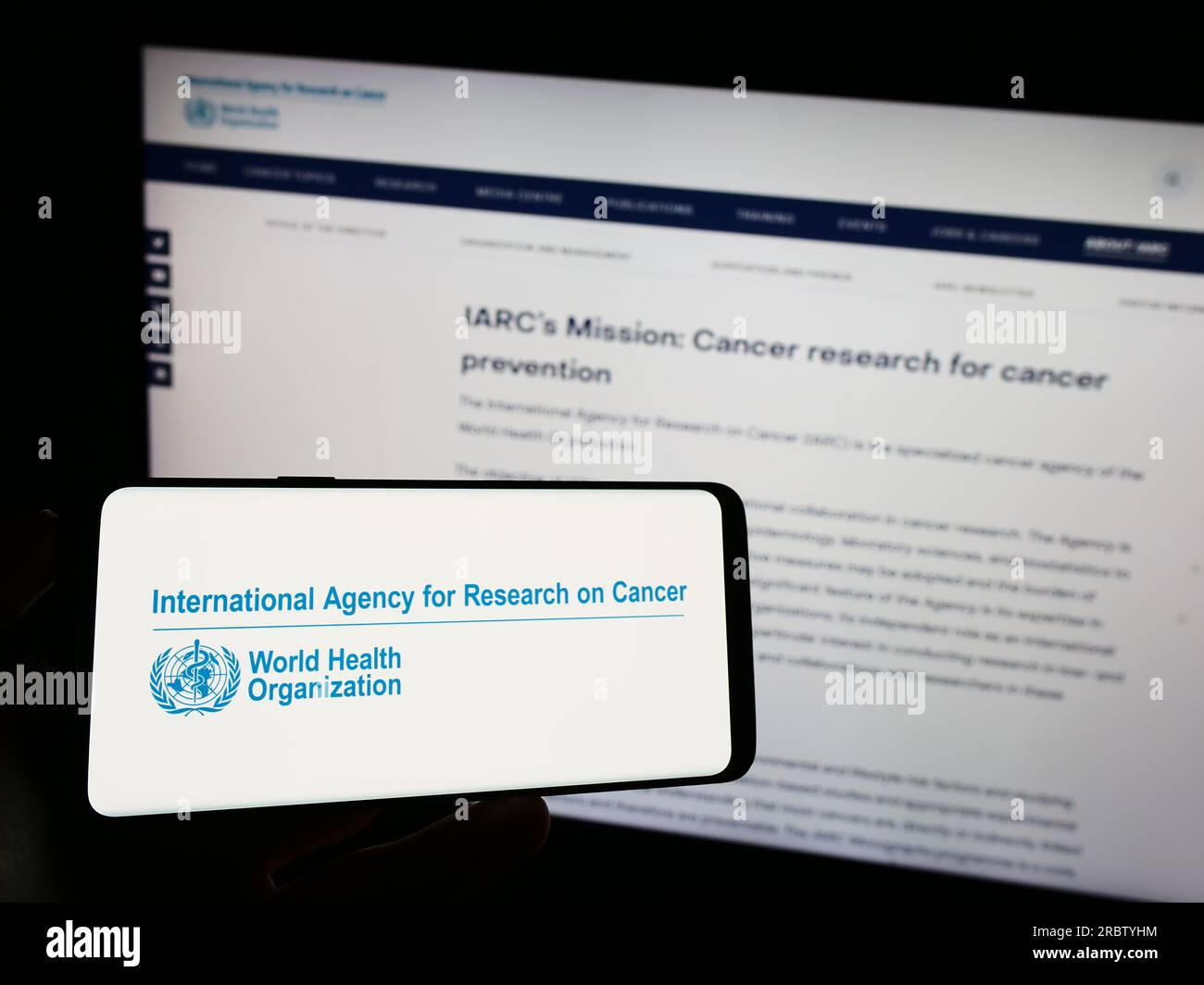 Persona che possiede un telefono cellulare con il logo dell'Agenzia internazionale per la ricerca sul cancro (IARC) sullo schermo davanti alla pagina web. Concentrarsi sul display del telefono. Foto Stock