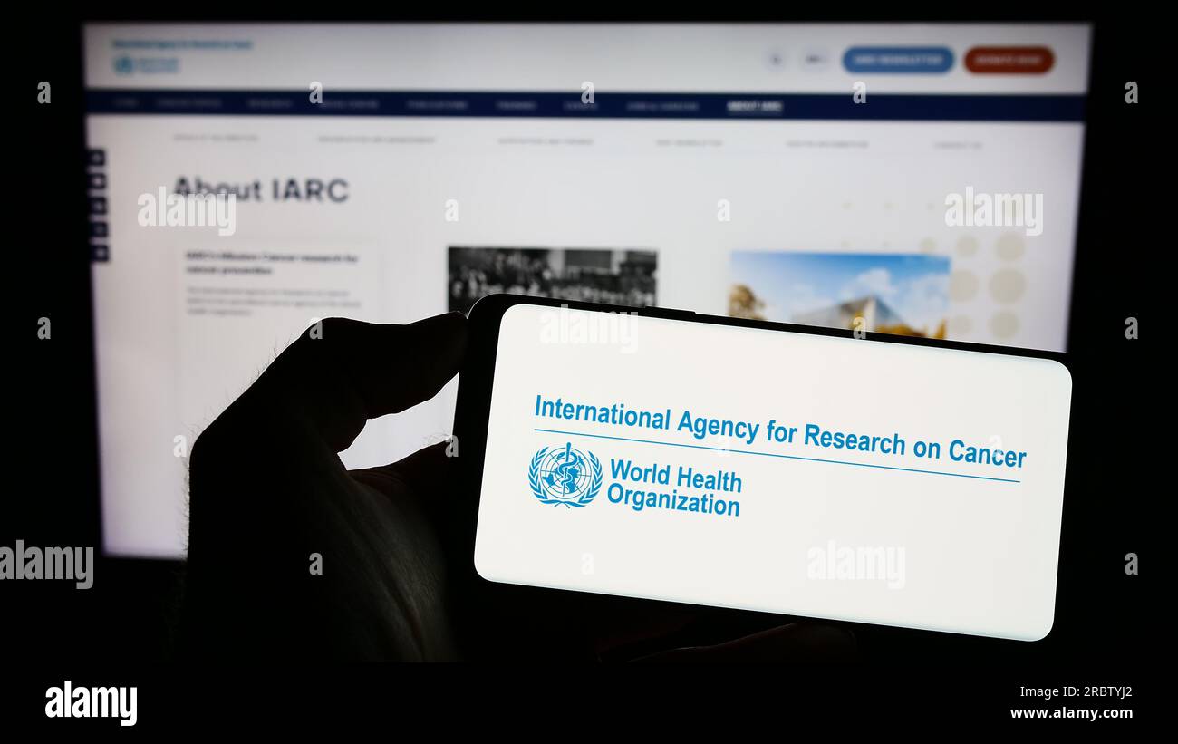 Persona che detiene il cellulare con il logo dell'Agenzia internazionale per la ricerca sul cancro (IARC) sullo schermo davanti alla pagina web. Concentrarsi sul display del telefono. Foto Stock