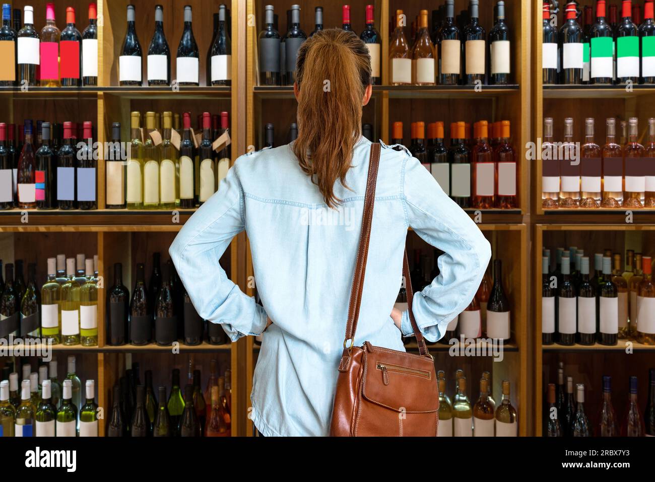 Foto retrovisiva della cliente che sceglie il vino in negozio, in piedi davanti agli scaffali con bottiglie di vino. Foto Stock