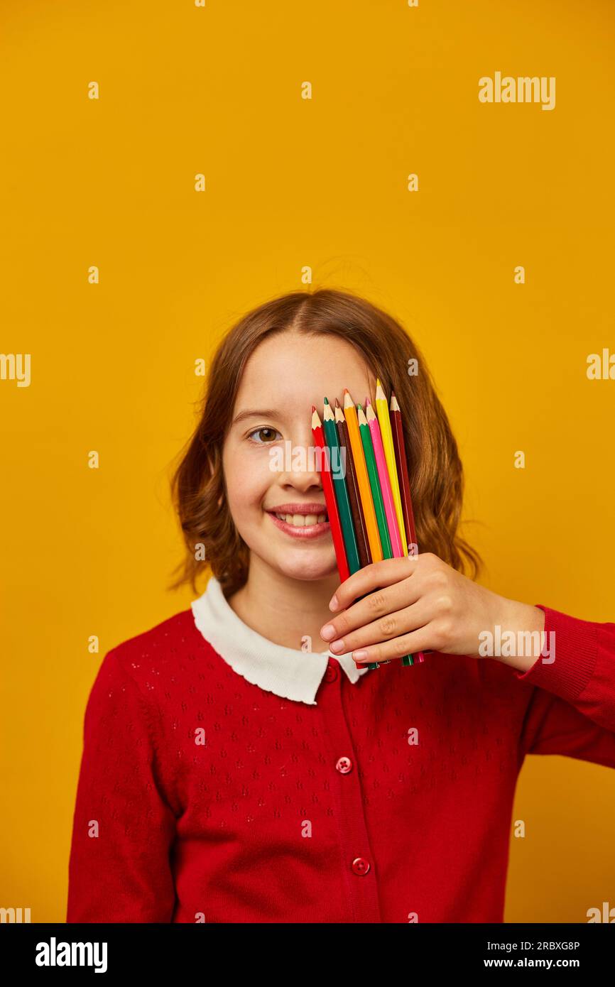 Ritrarre la ragazza adolescente che si copre il viso con matite colorate che si tengono in mano su sfondo giallo dello studio, concetto di ritorno a scuola, studio e.. Foto Stock