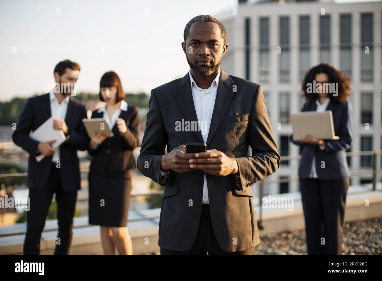 Ritratto di un imprenditore afroamericano di successo che tiene il telefono cellulare in mano e sorride davanti alla fotocamera. Partner commerciali di nazionalità mista che utilizzano Foto Stock