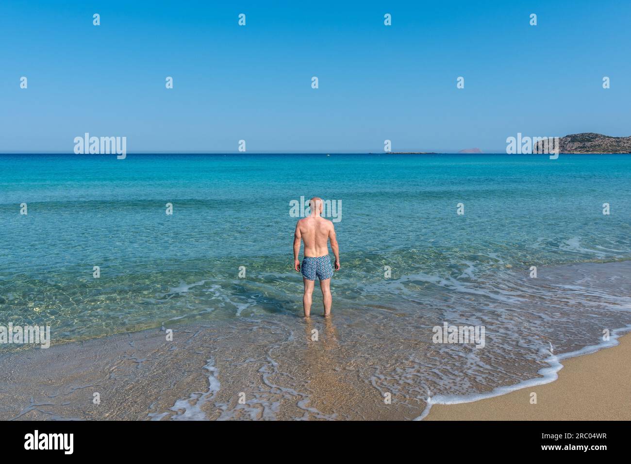 Vista dell'oceano turchese dalla spiaggia con un ragazzo in costume da bagno che guarda all'orizzonte. Desiderio di estate e libertà. Sabbia dorata. Foto Stock