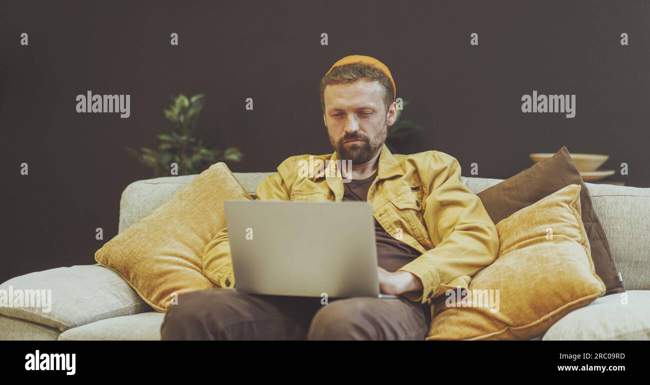 Uomo seduto sul divano con un notebook, impegnato in lavoro o in altre attività digitali. L'ambiente rilassato e confortevole del divano suggerisce che l'uomo si trovi in un ufficio domestico o in un ambiente interno accogliente. . Foto di alta qualità Foto Stock
