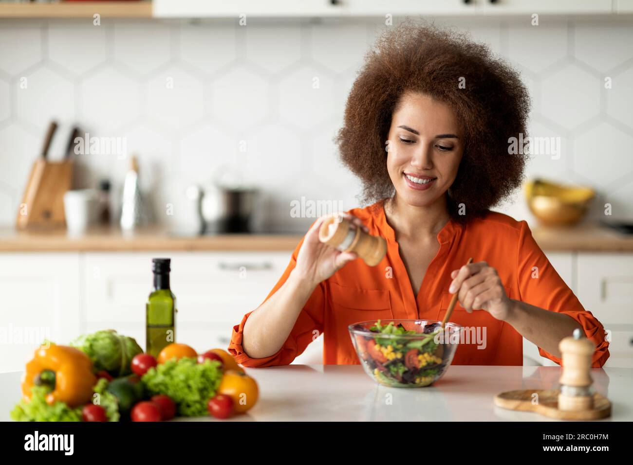 Felice giovane signora nera che prepara sana insalata di verdure in cucina, aggiungendo sale Foto Stock
