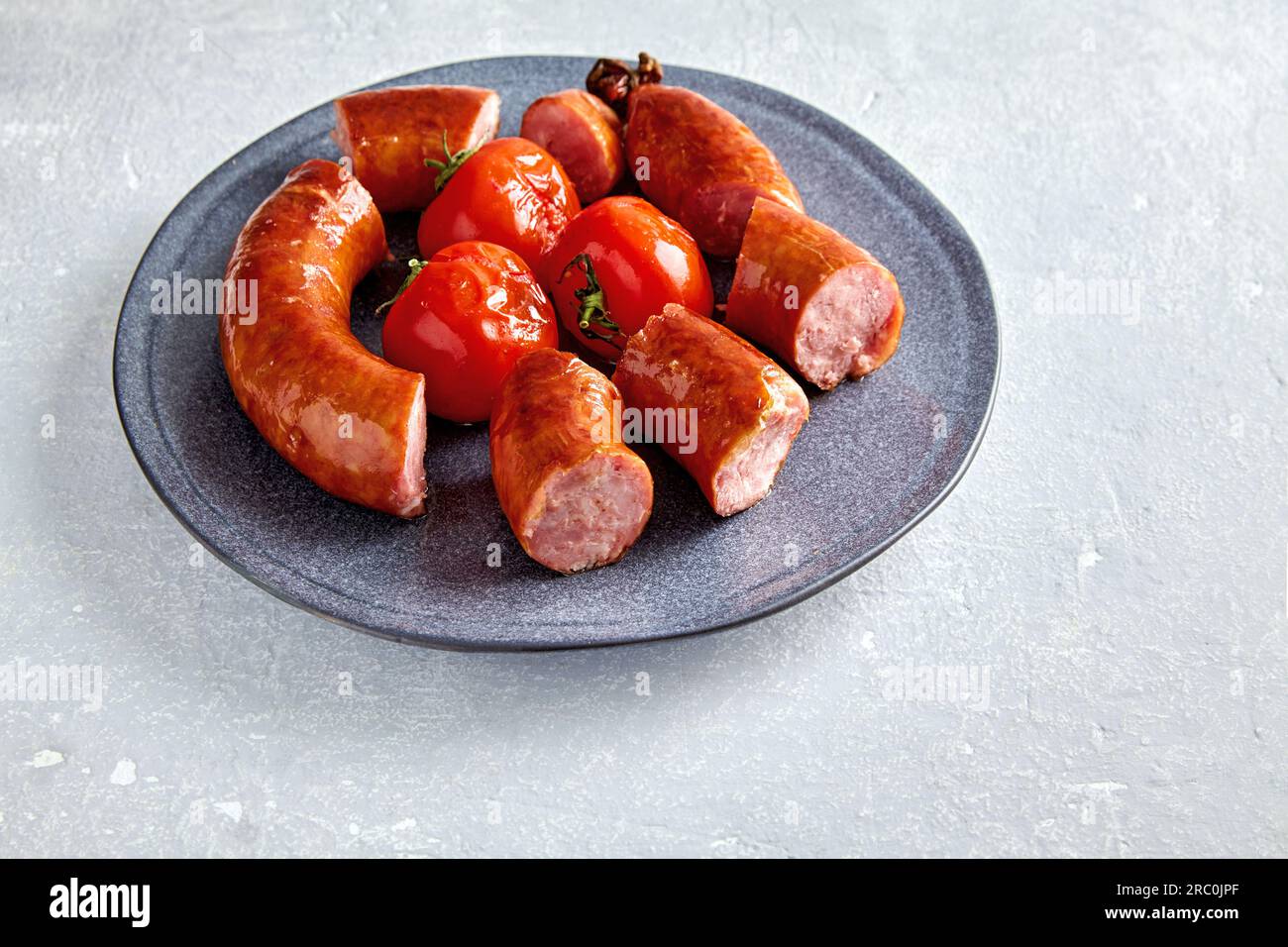 Salsiccia fritta a fette e pomodori grigliati su un piatto di ceramica grigia su una tavola di pietra leggera. Foto Stock