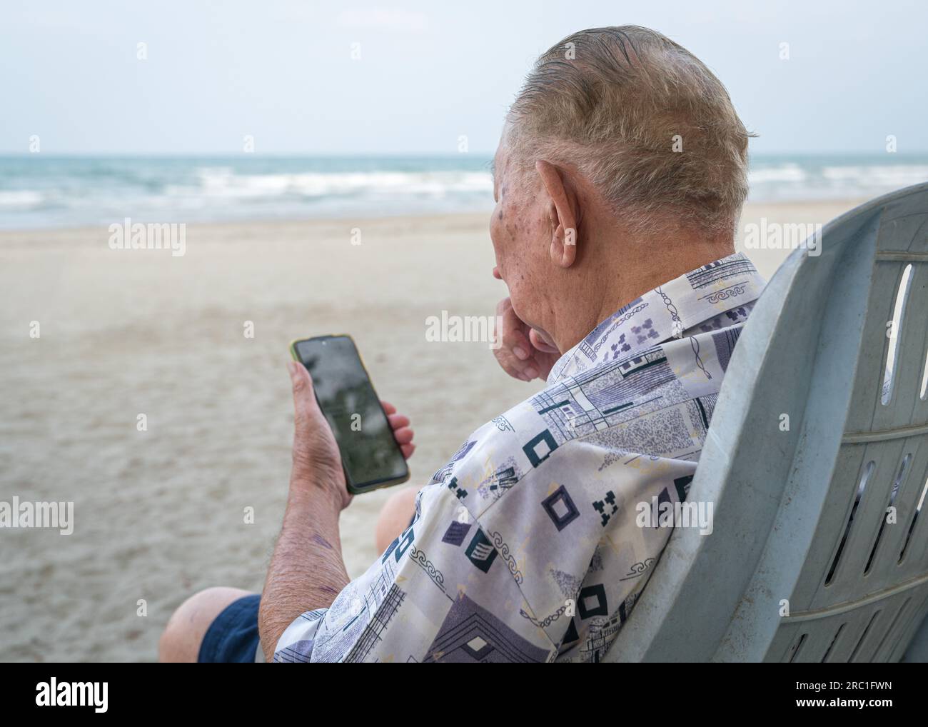 Uomo anziano seduto su una sedia a sdraio e che usa lo smartphone per leggere. Uso della tecnologia per gli anziani. Foto Stock