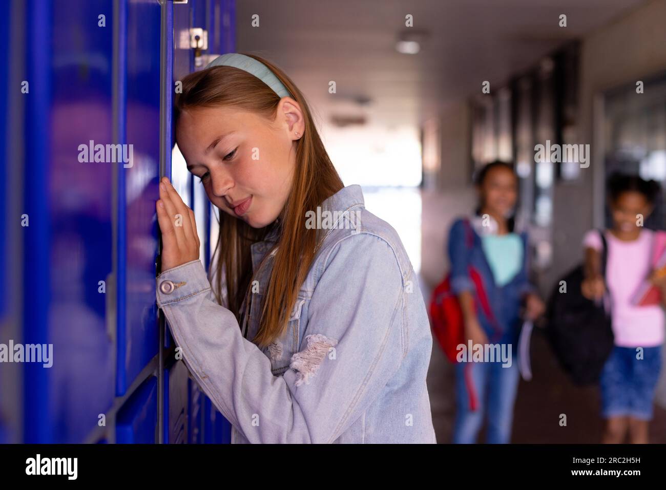 Triste studentessa caucasica in piedi accanto agli armadietti con diversi scolari sullo sfondo Foto Stock