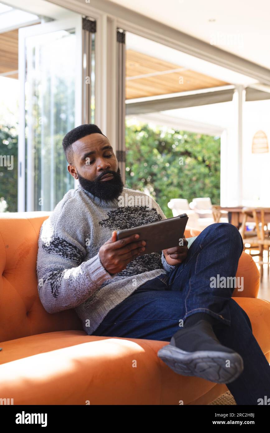 Uomo afroamericano concentrato che indossa maglie e usa un tablet sul divano a casa Foto Stock