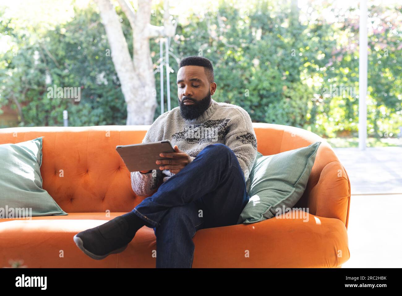Uomo afroamericano concentrato che indossa maglie e usa un tablet sul divano a casa Foto Stock