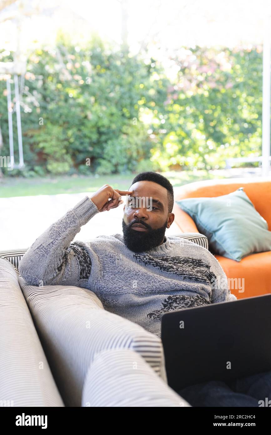 Un uomo afroamericano premuroso che indossa un maglione e usa un computer portatile sul divano a casa Foto Stock