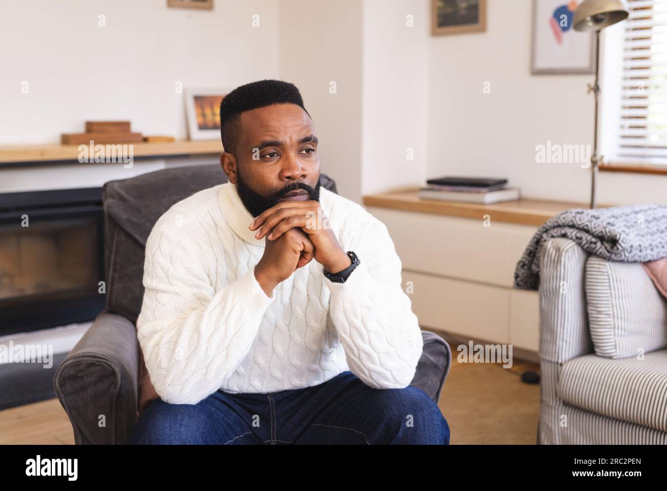 Un uomo afroamericano premuroso che indossa un maglione bianco e si siede su una poltrona a casa Foto Stock
