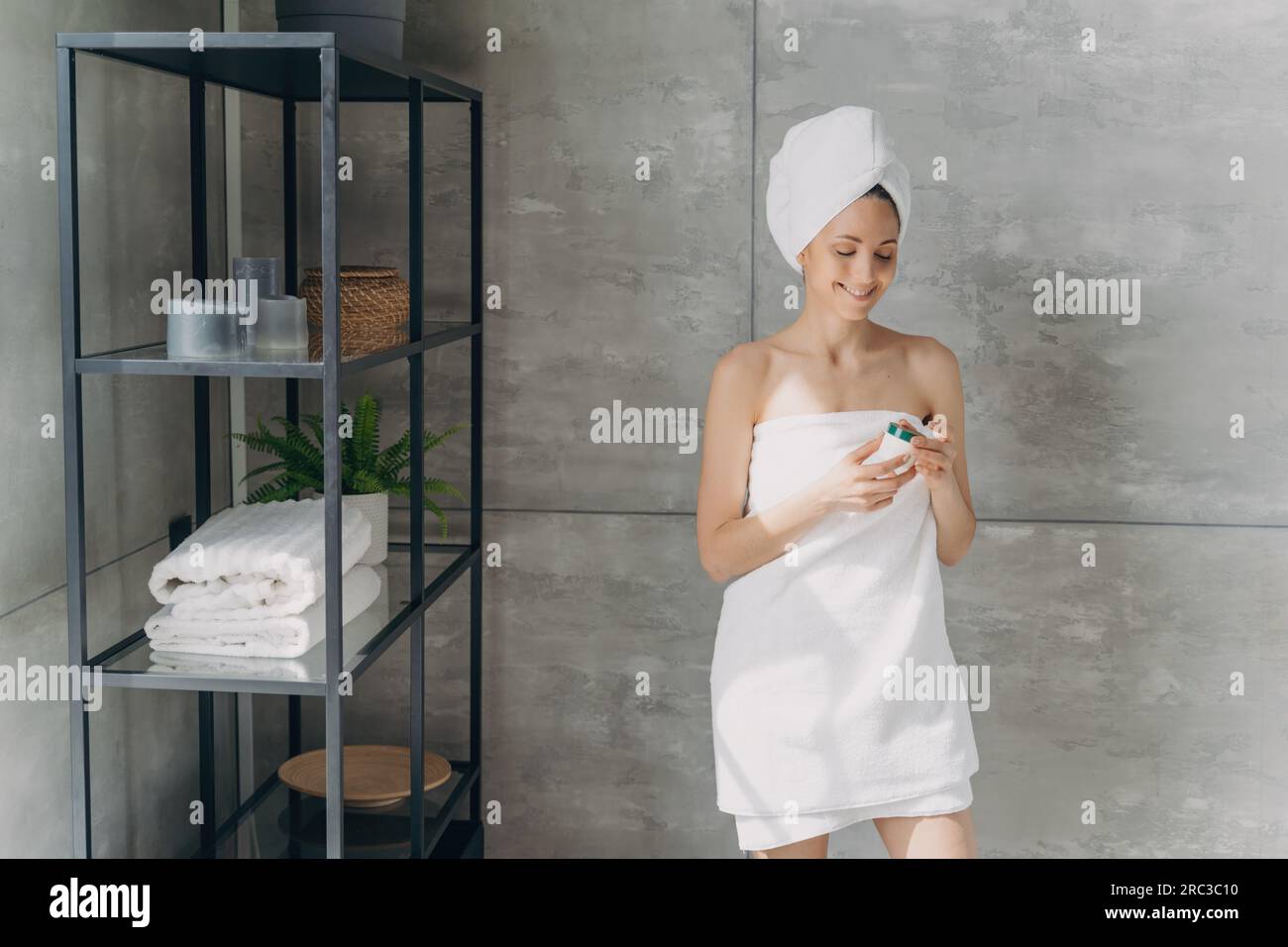La signora europea applica la crema dal barattolo. Bella ragazza in asciugamano dopo il bagno. La donna felice fa la doccia a casa. Concetto di cura della pelle. Foto Stock