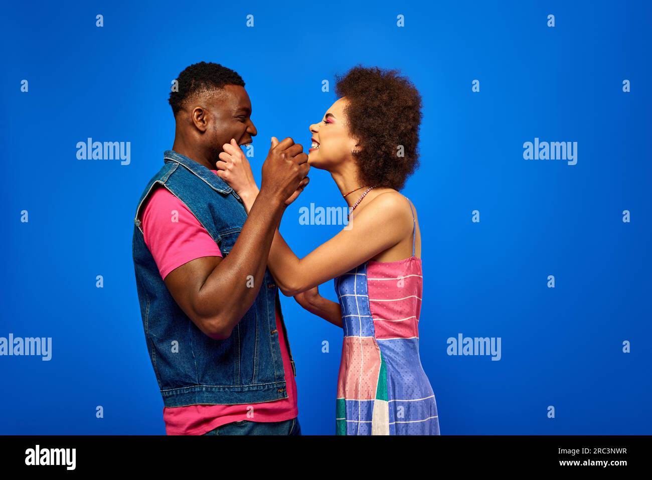 Vista laterale degli allegri amici afroamericani in luminosi abiti estivi che ridono mentre si alzano in piedi e si divertono insieme isolati su blu ed eleganti Foto Stock