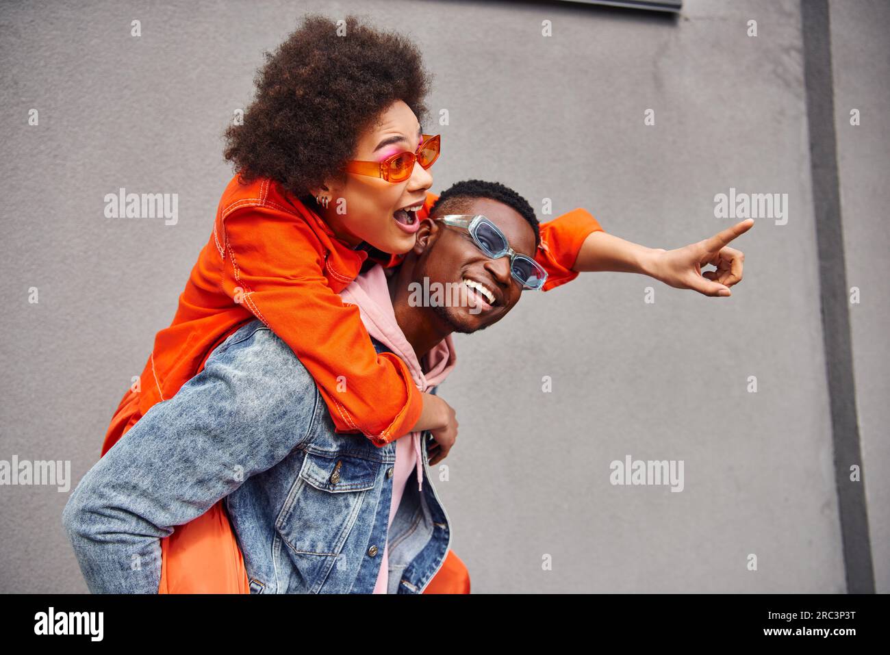 Eccitata giovane donna afro-americana con occhiali da sole ed elegante outfit che punta con il dito mentre si stringe con un amico e si trova vicino all'edificio Foto Stock