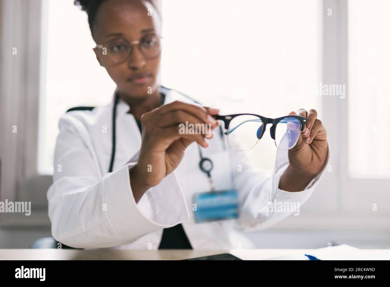 Medico afroamericano specializzato in colture con occhiali da vista, uniforme e stetoscopio che guarda in basso mentre si siede al tavolo e si controlla gli occhiali con il bl Foto Stock
