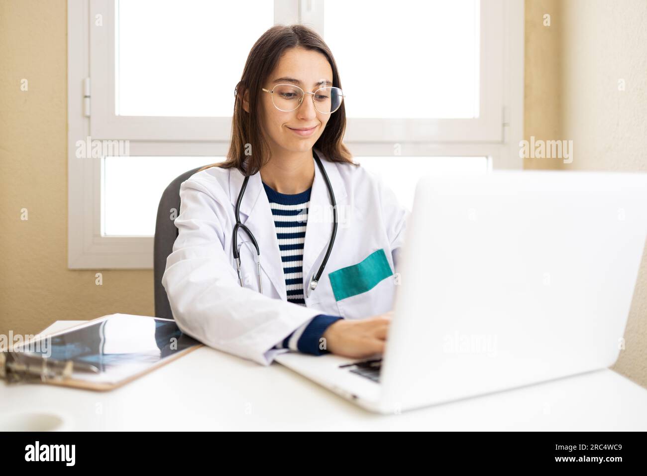 Giovane medico femminile in uniforme medica e occhiali seduti al tavolo con il computer portatile e dando consultazione online in ufficio Foto Stock