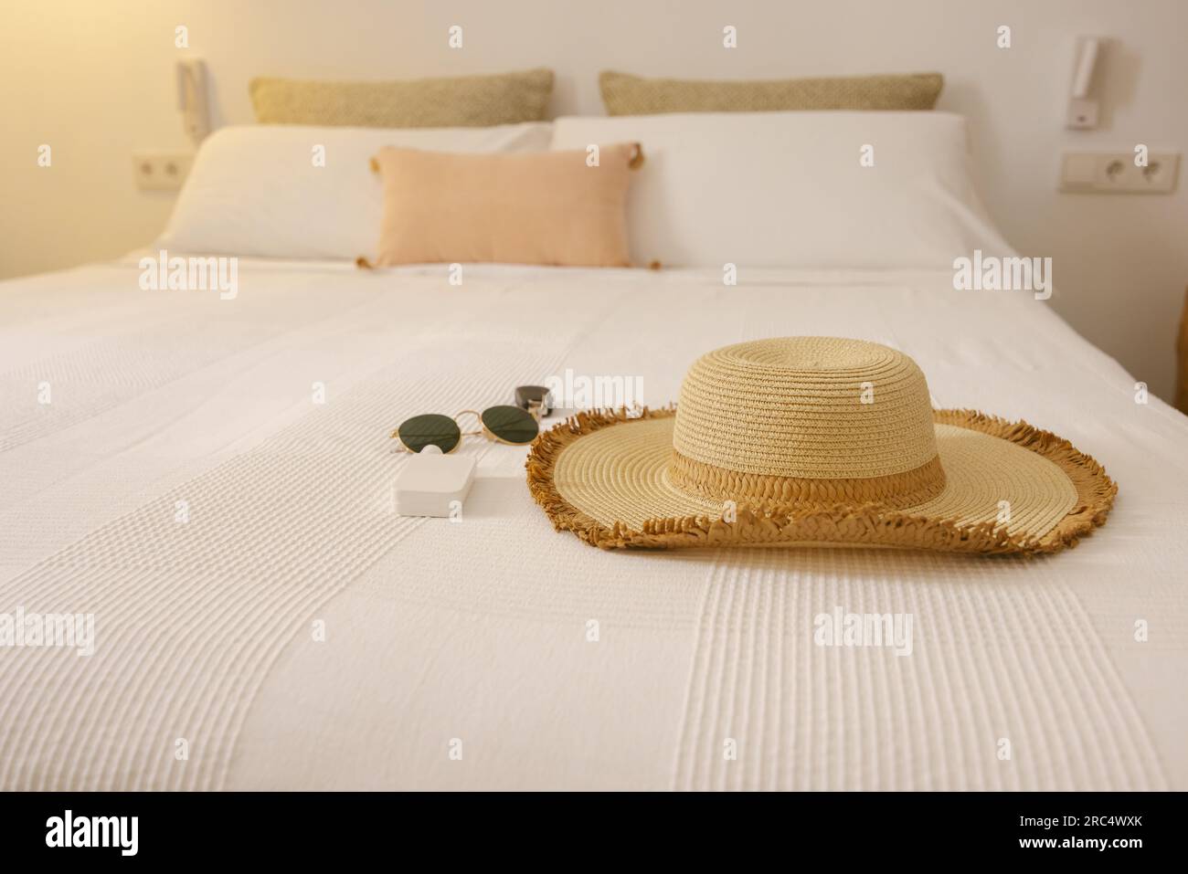 Cappello e occhiali da sole sul letto dell'hotel con cuscini bianchi e beige e coperta durante le vacanze estive nelle Isole Baleari Foto Stock