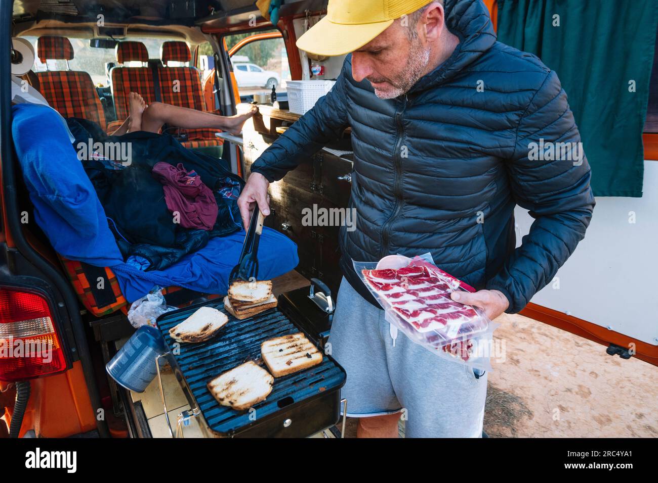 Viaggiatore maschile in giacca e berretto vicino al camper mentre tosta pane e carne alla griglia durante il viaggio nel canyon Rojo Teruel in Spagna Foto Stock