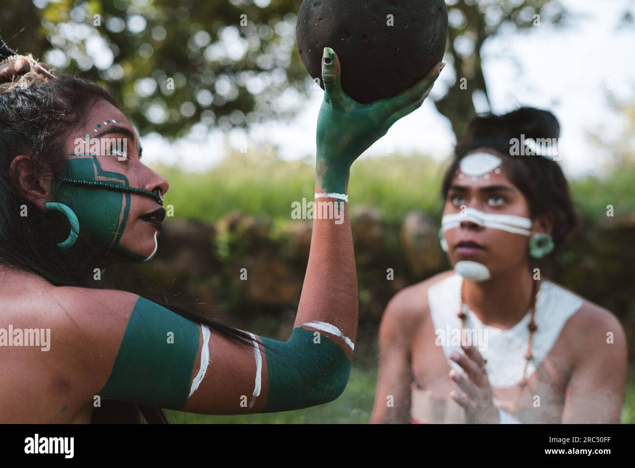 Vista laterale di giovani guerriere Maya etniche in abiti tradizionali con dipinti del corpo che guardano una palla di gomma nel parco verde Foto Stock