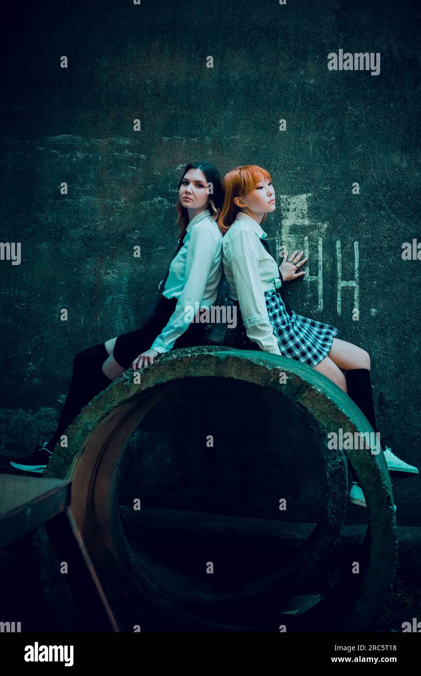In uno sfondo urbano di cemento industriale, due belle studentesse siedono fianco a fianco su un anello di cemento vicino a un muro. Foto Stock