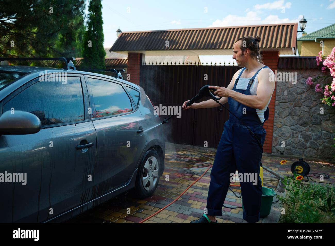 Bel muscoloso attraente giovane adulto in tute da lavoro blu, dirige un getto d'acqua ad alta pressione sulla sua auto mentre la pulisce all'aperto, utilizzando un Foto Stock
