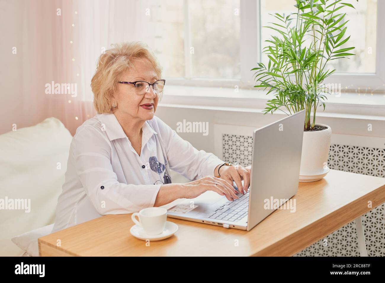 Ritratto di una persona affascinante e allegra sedersi dietro la scrivania con una tazza di caffè guardare interessato tv, lavorare da casa in interni. Felice donna matura che legge g Foto Stock