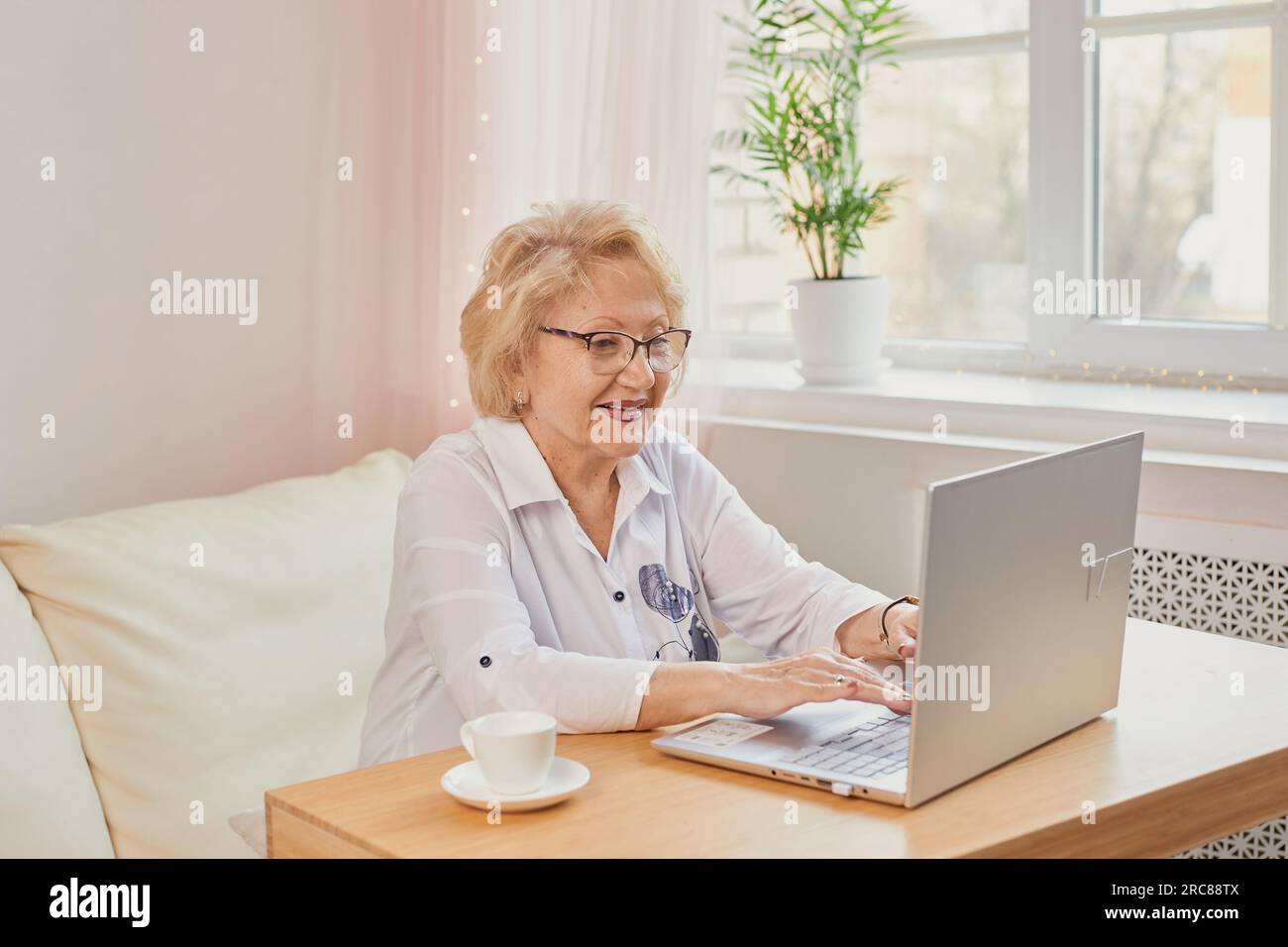Ritratto di una persona affascinante e allegra sedersi dietro la scrivania con una tazza di caffè guardare interessato tv, lavorare da casa in interni. Felice donna matura che legge g Foto Stock