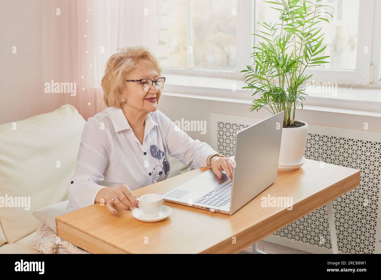 Ritratto di una persona affascinante e allegra sedersi dietro la scrivania con una tazza di caffè guardare interessato tv, lavorare da casa in interni. Felice donna matura che legge g Foto Stock