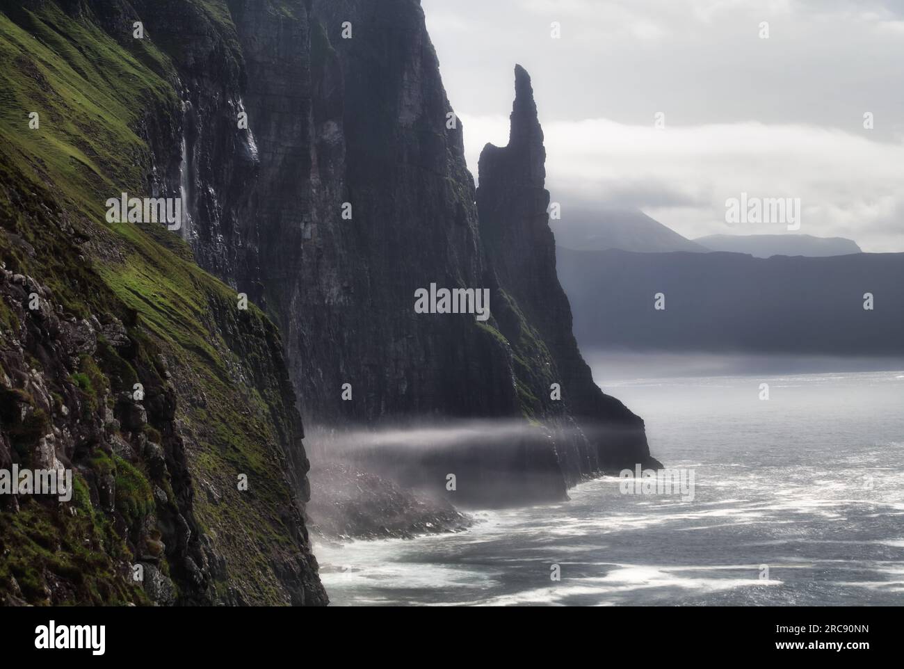 Costa rocciosa e Trøllkonufingur altrimenti nota come Troll Woman's Finger Over a Vagar, Isole Faroe nell'Oceano Atlantico. Foto Stock