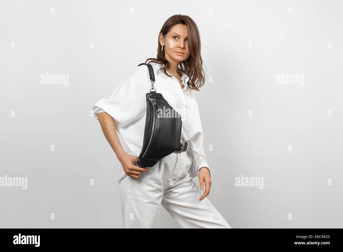 Donna bianca che indossa jeans bianchi, camicia e borsa a tracolla in pelle nera su sfondo grigio Foto Stock