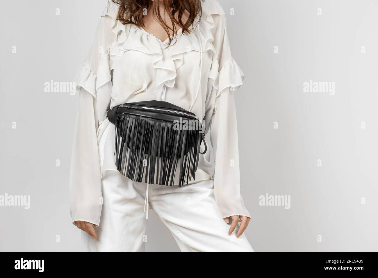 Donna bianca che indossa jeans bianchi, blusa con fronzoli e tracolla in pelle nera in vita con frangia decorativa su sfondo grigio Foto Stock