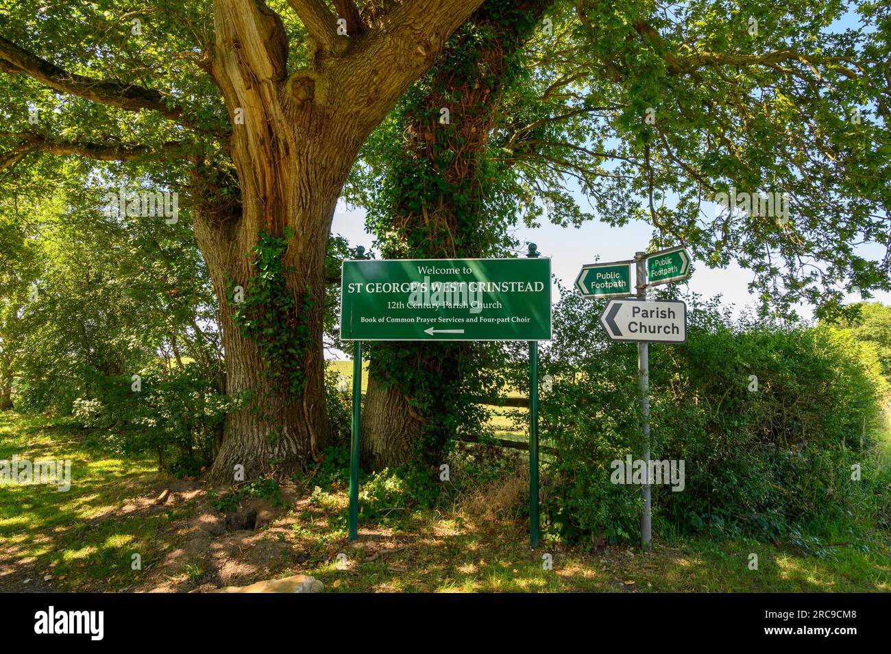 Cartelli con indicazioni per i sentieri e la chiesa parrocchiale West Grinstead di St George accanto a due querce mature. West Sussex, Inghilterra. Foto Stock