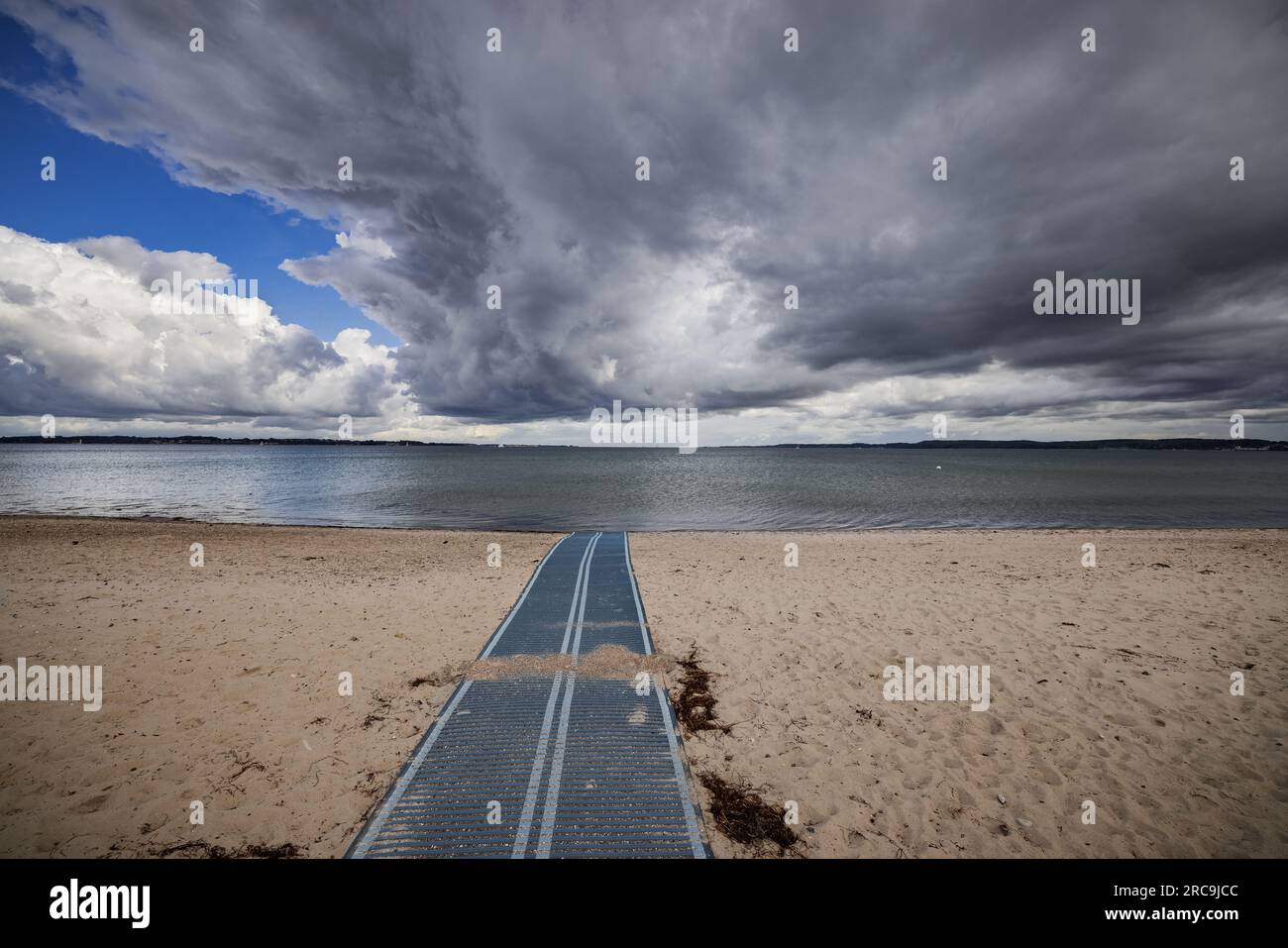 Dramatische Gewitterwolken über dem menschenleeren Strand von Holnis/Glücksburg. Eine Kunststoffbahn für Rollstuhlfahrer führt zum Flutsaum Foto Stock