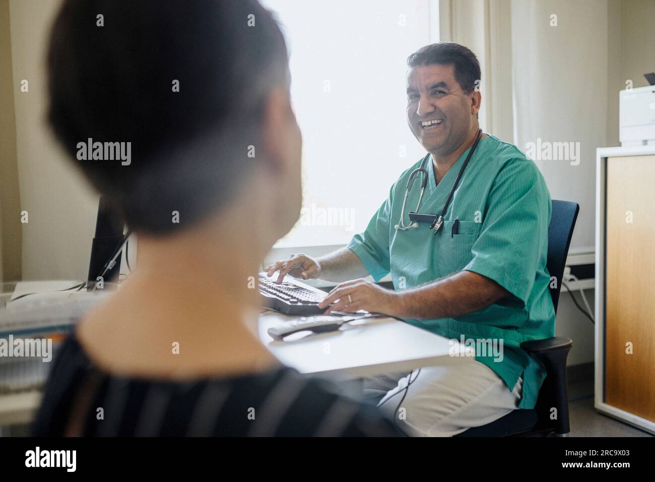 Medico maschio felice che guarda il paziente mentre usa il computer in clinica Foto Stock