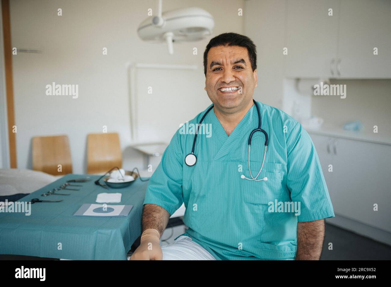 Ritratto di un medico maschio sorridente che indossa scrub seduto nella sala esame medico Foto Stock